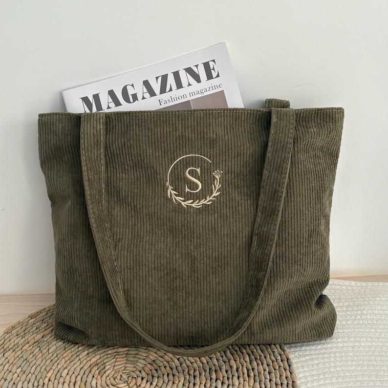 May include: Olive green corduroy tote bag with long handles. The bag features an embroidered floral wreath with the letter "S" in the center. A magazine with the word "MAGAZINE" is visible inside the bag.