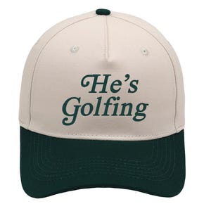 May include: A beige and dark green baseball cap featuring the text "He's Golfing". The cap has a curved peak and a button on top. The hat is a classic style, suitable for outdoor activities, with a dark green peak.