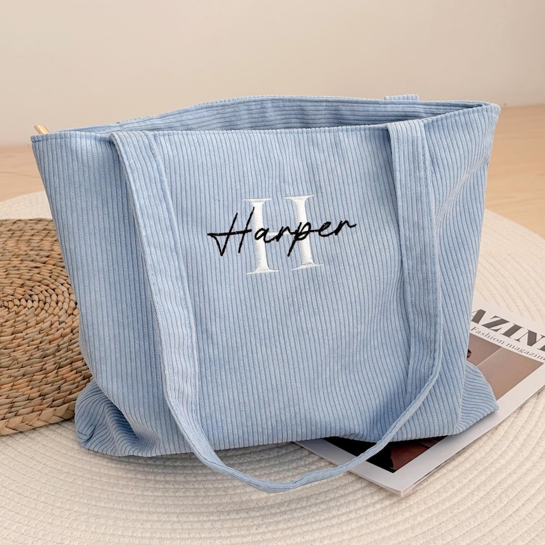 May include: A light blue corduroy tote bag with long handles. The word "Harper" is embroidered in black script, with the letter "H" and "II" in white. The bag is sitting on a white surface.