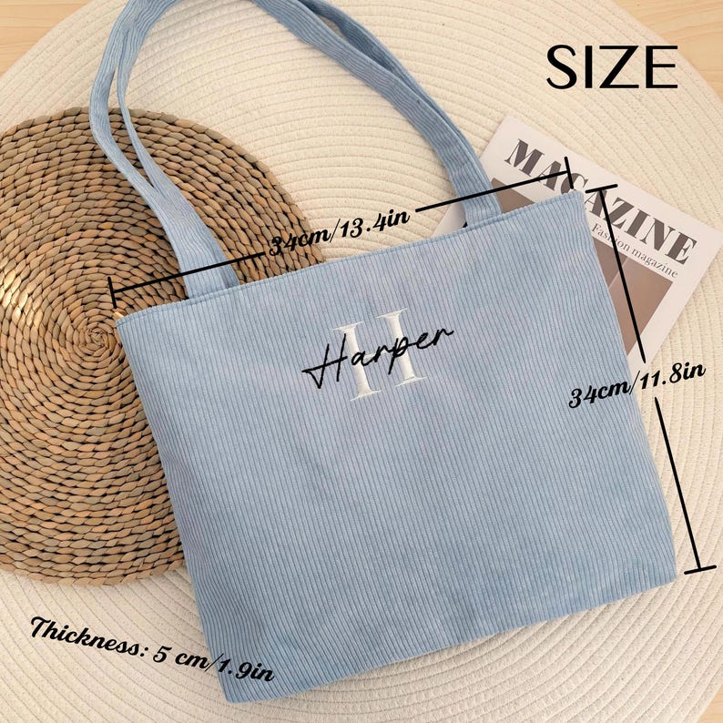May include: A light blue corduroy tote bag with the name "Harper" embroidered on the front. The bag is 13.4 inches wide and 11.8 inches tall, with a thickness of 1.9 inches. The bag has two handles and is sitting on a woven mat.
