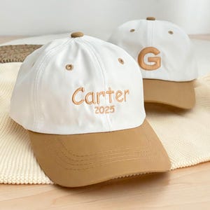 May include: Two white baseball caps with brown visors. One cap has the name "Carter" embroidered in gold with the year "2025" below it. The other cap has the letter "G" embroidered in gold. Both caps have a brown button on top.