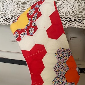 May include: A quilted Christmas stocking featuring a patchwork design of fabric hexagons. The pattern consists of solid red, solid yellow, and solid orange hexagons interspersed with white hexagons and floral-printed fabric in red, blue, and purple.