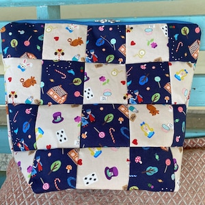 May include: A patchwork fabric zippered pouch featuring a whimsical Alice in Wonderland theme. The design incorporates squares of navy blue and beige fabric, adorned with illustrations of characters, sweets, and playing cards. The pouch has a blue zipper and trim.