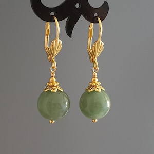 May include: A pair of gold-colored leverback earrings with a green, round stone. The earrings have a decorative gold-colored floral design at the base of the stone and at the top of the leverback.