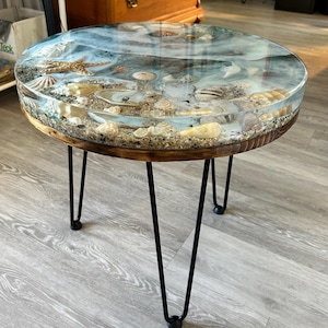 May include: Round side table with a resin top featuring seashells and sand, supported by black hairpin legs. The table's edge is made of wood. The table is approximately 24 inches in diameter.