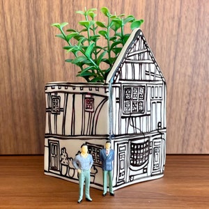 May include: A white ceramic planter shaped like a house with black line drawings of windows, doors, and a chimney. The planter has a small green plant growing out of the top and two miniature figurines standing in front of it.