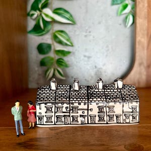 Puede incluir: Una miniatura de cerámica de una hilera de casas con detalles en blanco y negro, incluyendo un tejado de tejas y chimeneas. Dos figuras en miniatura están de pie frente a la casa. La casa se exhibe sobre una superficie de madera.