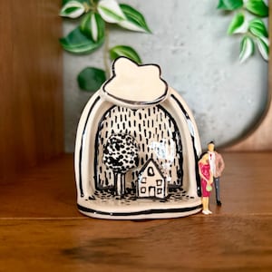 May include: A ceramic art piece featuring a black and white landscape scene with a tree, house, and rain-like pattern. The piece has a cream-colored exterior with black trim and a small figurine of a couple.