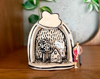 May include: A cream-colored ceramic arch sculpture featuring a black hand-painted illustration of a house and a tree against a rain-like background. A miniature figurine of a couple stands to the right of the arch, which has a cloud-shaped top.