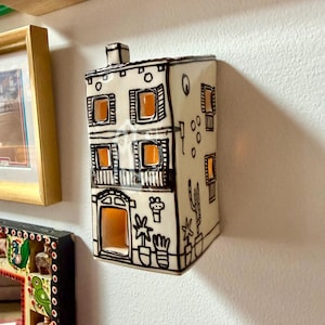 Może przedstawiać: Ścienny świecznik ceramiczny w kształcie domu w kolorze kremowym z czarnymi, ręcznie malowanymi detalami liniowymi. Konstrukcja posiada wycięte okna i drzwi, ukazujące pomarańczową poświatę wnętrza, ozdobiona ilustracjami roślin w doniczkach.