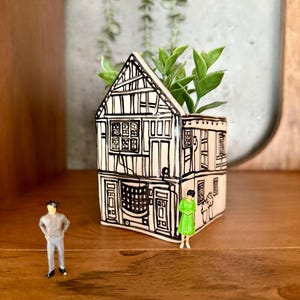 Può includere: Un vaso di ceramica a forma di casa, con dettagli architettonici in bianco e nero. Contiene piante verdi. Miniature di un uomo e una donna sono nelle vicinanze. Il vaso è su una mensola di legno.