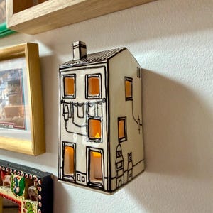 Op de afbeelding: Een keramische wandsculptuur in de vorm van een huis in off-white met zwarte handgetekende contouren. Het object heeft uitgesneden ramen met een warme interne gloed, een schoorsteen en gedetailleerde illustraties van was aan een lijn.