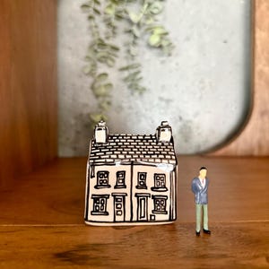 Peut inclure: Une petite maison en céramique avec un toit et des bordures noirs et blancs, avec plusieurs fenêtres et deux cheminées. Une figurine d'une personne en costume se tient à côté de la maison. La maison est exposée sur une surface en bois.