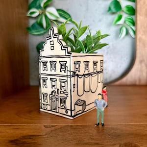 Puede incluir: Una maceta beige con forma de casa con detalles de contorno negro, llena de plantas verdes. La maceta tiene un diseño de fachada detallado. Figuras en miniatura de un hombre y una mujer están cerca. La maceta está sobre una superficie de madera.