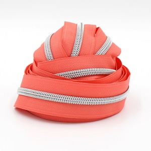 May include: A close-up of a roll of coral colored zipper tape with a silver zipper teeth. The zipper tape is coiled up and ready to be used for sewing projects.