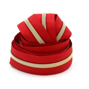 May include: Red zipper tape with a gold metal zipper pull. The tape is coiled and ready to be used for sewing projects.
