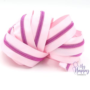 May include: Pink zipper tape with a purple zipper teeth. The tape is coiled and ready to be used for sewing projects.