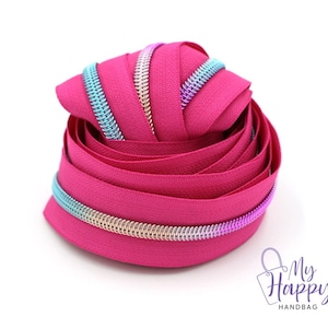 May include: A close-up of a pink zipper tape with a rainbow colored zipper pull. The zipper tape is coiled up and the zipper pull is visible at the bottom of the image.