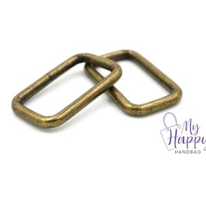 May include: Two antique gold metal D-rings, also known as D rings or D-shaped rings, are shown on a white background. These rings are commonly used in crafting, sewing, and leatherwork.