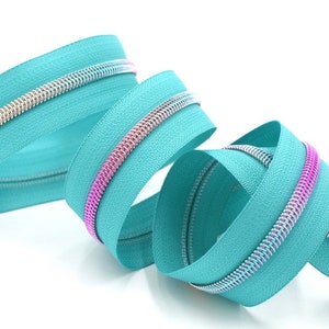 May include: A close-up of a turquoise zipper with a silver and iridescent pink zipper tape. The zipper is coiled and ready for sewing.