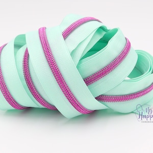 May include: A close-up of a light green zipper with a pink zipper tape. The zipper is coiled and ready to be used for sewing projects.
