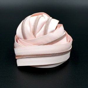 May include: A close-up of a pink zipper tape with a rose gold zipper pull. The zipper tape is coiled up and the zipper pull is visible in the center of the coil.