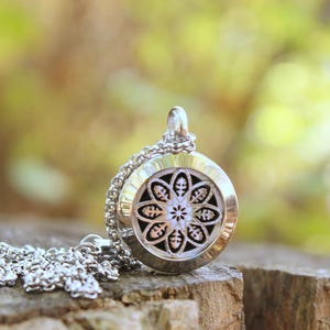 May include: Silver-toned pendant necklace with a detailed floral design. The round pendant hangs from a delicate chain and rests on a wooden surface. The background is a soft, blurred mix of green and brown.