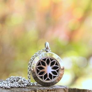 May include: A silver-colored pendant necklace with a floral design. The pendant has a circular shape with a black interior and a flower-like pattern. The necklace is displayed on a wooden surface against a blurred green and yellow background.