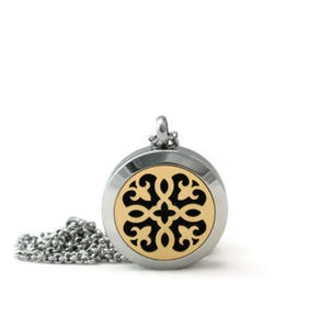 May include: A circular silver-toned metal pendant necklace featuring a gold-toned center inlay with a black cutout filigree pattern. The pendant is attached to a silver-toned metal chain that is coiled on the side, creating a polished accessory.