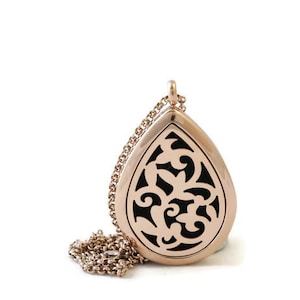 May include: A rose gold teardrop-shaped locket pendant featuring an intricate cutout filigree pattern over a solid black background. The pendant is attached to a matching rose gold link chain that rests in a coiled pile beside the locket.