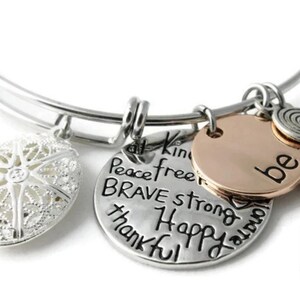May include: A silver-toned expandable bangle bracelet featuring three charms: a filigree oval charm, a round silver charm engraved with the words Peace, free, BRAVE, strong, thankful, Happy, and a round rose gold charm engraved with the word be.
