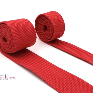May include: Two rolls of bright red webbing material, unspooled, against a white background. The material is tightly woven and appears to be made of a durable fabric. The rolls are positioned diagonally, showcasing the texture and color.