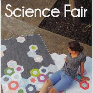 May include: A woman sits on a colorful hexagonal quilt with a gray hexagonal quilt behind her. The quilts are on a concrete surface. The text "Science Fair" is at the top of the image. The text "Jaybird Quilts" is at the bottom of the image.