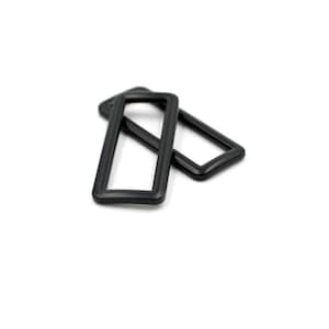 Puede incluir: Dos anillos rectangulares negros de plástico se superponen ligeramente sobre un fondo blanco. Los anillos tienen esquinas redondeadas y un acabado liso y mate. La imagen está bien iluminada, destacando la forma y textura de los objetos.