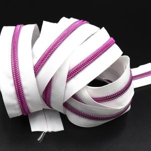 Puede incluir: Cinta de cremallera blanca con una bobina metálica rosa. La cinta está enrollada y doblada.