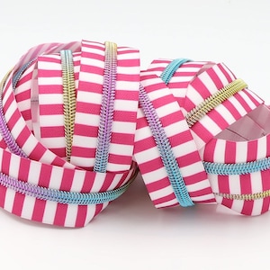 Może przedstawiać: Zbliżenie na taśmę suwaka w paski różowe i białe z tęczowymi zębami suwaka. Taśma jest zwinięta, a zęby suwaka są widoczne.