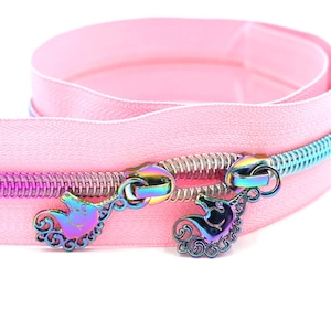May include: Pink zipper tape with iridescent metal zipper pulls shaped like unicorns. The zipper tape is folded in half, showing the inside of the tape.