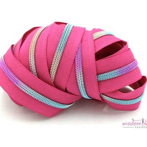 May include: A close-up of a pink zipper tape with a silver and iridescent blue zipper coil. The zipper tape is coiled and folded on a white surface.