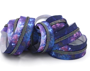 Puede incluir: Primer plano de una pila de cinta de cremallera morada y azul con estampado de galaxia. La cinta de cremallera tiene una cremallera plateada.