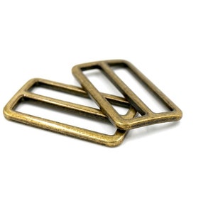 May include: Two antique gold metal slider buckles. The buckles are rectangular with a smooth finish and a slightly rounded edge.