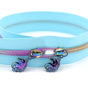 Peut inclure: Une fermeture éclair bleu clair avec un curseur doré et iridescent. Le curseur est orné de deux breloques en forme de licorne.