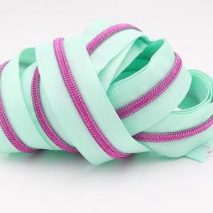 May include: A close-up of a light green zipper with a pink zipper tape. The zipper is coiled up and ready to be used for sewing projects.