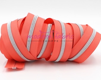 May include: A close-up of a roll of coral orange zipper tape with a silver zipper. The zipper tape is made of nylon and the zipper is made of metal.