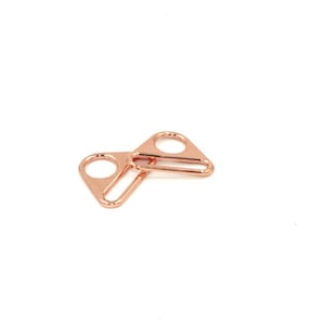 Può includere: Due anelli triangolari in metallo oro rosa con apertura circolare e barra rettangolare. L'hardware è progettato per l'uso nella fabbricazione di borse, cucito o altri progetti artigianali. Gli anelli sono esposti su sfondo bianco.