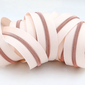 May include: A close-up of a white zipper with a rose gold zipper pull. The zipper is coiled up and ready to be used for sewing projects.