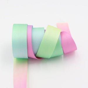 May include: A roll of woven ribbon featuring a gradient color pattern that transitions through shades of mint green, soft pink, light blue, and pale yellow. The ribbon has a smooth, consistent texture and is coiled into a cylindrical shape.