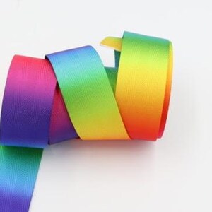 May include: A rainbow ombre ribbon with a herringbone weave. The ribbon is rolled up and the colors are red, orange, yellow, green, blue, indigo, and violet.