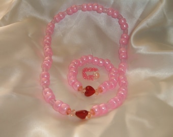Könnte beinhalten: Ein rosafarbenes Perlen-Halsketten- und Armband-Set mit roten Herz-Anhängern. Die Halskette und das Armband bestehen aus kleinen, runden Perlen.
