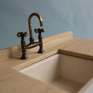 以下が含まれることがあります： 木製のミニチュアキッチンシンク。白い磁器製のシンクと真鍮製の蛇口が2つのハンドルで備わっています。
