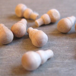 Könnte beinhalten: Eine Ansammlung kleiner, unfertiger Holzknebel, jeder mit einer abgerundeten Oberseite und einem zylindrischen Sockel. Die Knöpfe sind auf einer grauen Oberfläche verstreut und zeigen ihre natürliche Holzmaserung und ihr schlichtes Design.